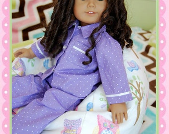 May include: A doll with long, dark curly hair wearing purple polka dot pajamas with white trim. The doll is seated on a white and cream-colored beanbag chair decorated with colorful owl and tree designs.