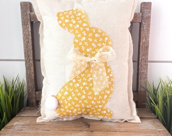 May include: A rectangular cream-colored pillow with a yellow floral bunny applique and a sheer bow. The bunny has a white pom-pom tail. The pillow is placed on a rustic wooden bench, suggesting a spring or Easter theme.