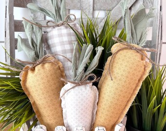May include: A decorative Easter basket with three fabric carrots. The carrots are yellow, white, and tan, with green fabric tops and tied with twine. The basket is white and has a picket fence design.