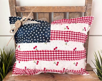 May include: A decorative rectangular pillow with a patriotic theme. The pillow features a navy blue section with white stars, red and white gingham stripes, and a white fabric with red cherries. It is adorned with a button and twine.