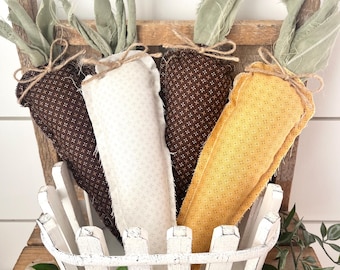 May include: A rustic white picket fence basket holds four fabric carrots. The carrots are brown, white, and yellow, with green fabric tops tied with twine. The basket sits on a wooden surface with greenery.