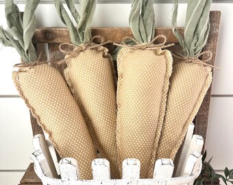 May include: Four fabric carrots with green fabric tops and tan polka-dot bodies are displayed in a white picket fence basket. The basket sits on a wooden surface, with greenery adding a natural touch. The carrots are tied with twine.