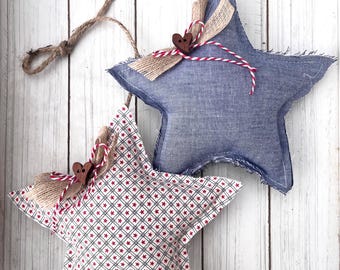 May include: Two star-shaped ornaments. One is denim blue, the other is patterned with red stars on a white background. Both have burlap accents, wooden heart-shaped buttons, and red and white twine. They hang from a natural rope.