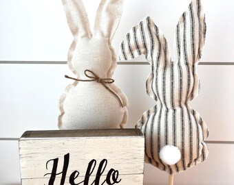 May include: Two decorative fabric bunny figures and a wooden block sign that reads "Hello Spring". One bunny is solid beige, the other is striped. The sign is white with black lettering. A string of white felt balls hangs in the background.