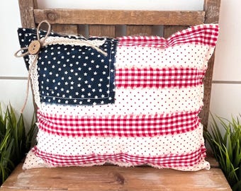 May include: A decorative pillow with a patriotic theme, resembling the American flag. The pillow features a navy blue square with white stars, red and white gingham stripes, and white polka dots. It is adorned with a wooden button and twine.