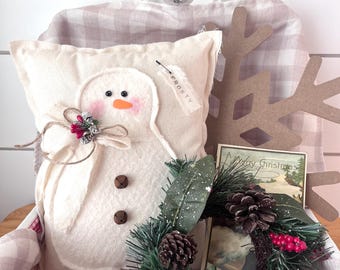 May include: A festive Christmas display featuring a snowman pillow with a bow, a small wreath with pinecones, and a vintage Christmas card. The pillow has a tag that says "Frosty". The items are arranged in a plaid basket.