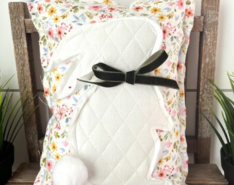 May include: A decorative pillow featuring a white quilted bunny design with a fluffy tail and a dark green bow. The pillow has a floral pattern with pink, yellow, and blue flowers on a white background. The pillow is sitting on a wooden chair.