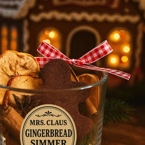 May include: A clear glass jar filled with dried apple slices, cinnamon sticks, and a gingerbread man. The jar has a label that reads "MRS. CLAUS GINGERBREAD SIMMER POT" and is tied with a red and white checkered ribbon. A gingerbread house is in the background.