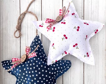 May include: Two star-shaped fabric ornaments. One is navy blue with white stars, the other is white with red cherries. Both have a jute string for hanging, a wooden button, and a red and white gingham bow. Decorative home decor.