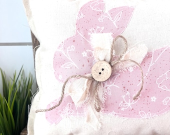 May include: A decorative pillow with a pink bunny applique. The bunny is made of pink fabric with a white floral pattern. A wooden button and twine bow are attached to the bunny. The pillow is on a wooden surface.