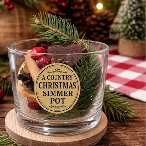 May include: A clear glass jar filled with Christmas simmer pot ingredients, including cranberries, cinnamon sticks, and orange slices. The jar has a gold label that reads "A Country Christmas Simmer Pot." The jar sits on a wooden coaster, with pine branches and a small Christmas tree in the background.
