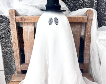 May include: A white ghost figurine draped in sheer, textured fabric, featuring two oval-shaped black mesh eyes and wearing a small, pointed black felt witch hat. The ghost is positioned upright, with the fabric cascading down in loose, frayed folds.