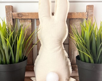 May include: A cream-colored fabric bunny with a fluffy white tail sits on a wooden surface. The bunny is flanked by two black potted plants with green grass. The background features a rustic wooden structure and a white wall.