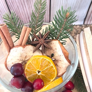 May include: A clear glass bowl filled with dried apple slices, a sliced orange, cranberries, star anise, cinnamon sticks, and evergreen sprigs. The arrangement is set against a light wooden background, creating a festive and aromatic display.