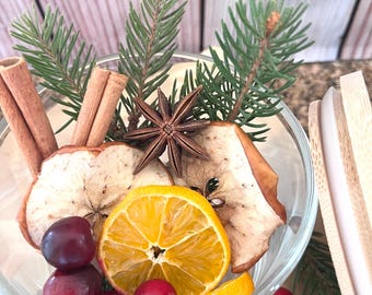 May include: A clear glass bowl filled with dried apple slices, a sliced orange, cranberries, star anise, cinnamon sticks, and evergreen sprigs. The arrangement is set against a light wooden background, creating a festive and aromatic display.