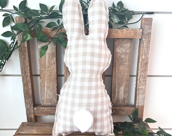 May include: A beige and white gingham bunny-shaped pillow with a white pom-pom tail. The decorative pillow is placed on a rustic wooden chair, with green leafy vines in the background. The bunny is a seasonal Easter or spring decoration.