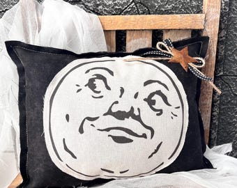 May include: A rectangular black fabric pillow featuring a large, centered white circular graphic of a moon with a human-like face. The pillow is accented with a small, rusted metal star tied to the top corner with black and white twine.