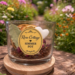 May include: A clear glass simmer pot with a wooden lid, filled with dried botanicals, a white heart, and a slice of dried orange. The label reads "Rose Cottage Simmer Pot". The pot sits on a wooden surface with a blurred garden background.