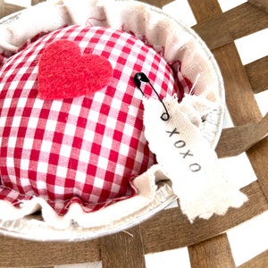 May include: A red and white gingham fabric pie crust pincushion with a red felt heart and a safety pin. The pincushion is sitting in a brown wicker basket with a white fabric tag that says "XOXO".