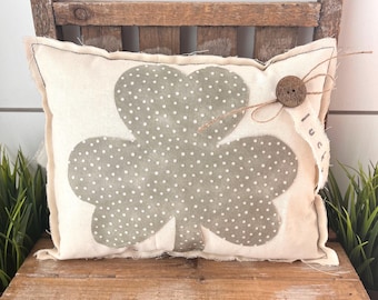 May include: Cream-colored pillow with a large gray shamrock design and white polka dots. A brown button and twine accent the pillow's edge, with a small fabric tag that reads "luck". The pillow is placed on a rustic wooden chair.
