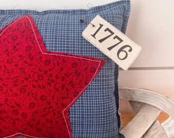 May include: A decorative pillow with a red star applique on a blue plaid background. The star has a floral pattern and white stitching. A tag with the number "1776" is attached to the pillow.