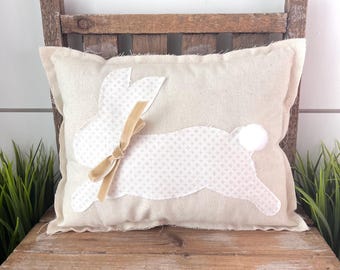 May include: A rectangular beige pillow with a white bunny applique. The bunny has a patterned body, a tan velvet bow, and a white pom-pom tail. The pillow is placed on a rustic wooden chair, with green grass on either side.