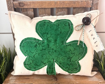 May include: A cream-colored pillow with a large green shamrock applique. The shamrock has a swirling pattern and is stitched onto the pillow. A small tag that says "lucky" is attached with twine and a bell.