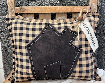 May include: A square throw pillow featuring a black and tan buffalo check pattern. A black fabric house silhouette is appliqued in the center. A small white fabric tag with the word haunted is tied to the top corner with twine and a bell.