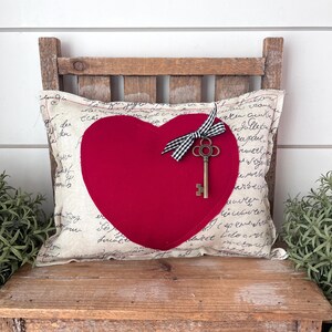 May include: A decorative pillow with a large red heart applique. The pillow has a cream-colored background with script writing and is adorned with a vintage key and a black and white checkered bow. The pillow is on a wooden chair.