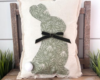 May include: A rectangular pillow with a light-colored fabric cover. A green bunny silhouette with a floral pattern is centered on the pillow. A black bow is tied around the bunny's neck, and a white pom-pom tail is attached. The pillow is placed on a wooden surface.
