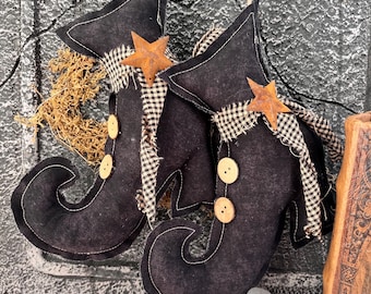 May include: Pair of black witch boots with a rustic star accent and gingham trim. The boots have wooden buttons and are suspended by twine. A decorative Halloween or fall-themed item.