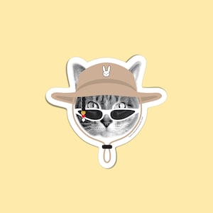 Op de afbeelding: Een gestanste sticker met een zwart-witte halftoonillustratie van een kat die een beige vissershoed draagt met een klein wit konijntje, een witte cat-eye zonnebril en een enkele gevlochten haarlok met een rood hartje en witte kraal.