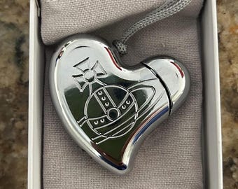 May include: A heart-shaped, silver-colored pendant with a detailed emblem. The pendant is attached to a silver cord and rests inside a white box with a gray fabric lining. The emblem features a cross and a planet-like design.