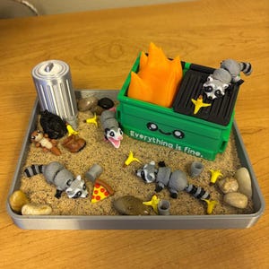 May include: A miniature diorama featuring a green dumpster with orange flames, raccoons, and an opossum. The scene includes a trash can, pizza slice, and the text "Everything is fine." The setting is a tray with sand and small rocks.