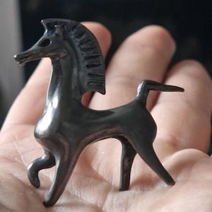 May include: A small, dark gray horse figurine with a stylized design. The horse has a long neck, a flowing mane, and a raised tail. The figurine is held in a person's hand, and the background is blurred.
