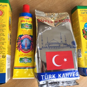 Op de afbeelding: Drie gele en rode pakjes specerijen. Het eerste pakje is een tube rode peperpasta met het label "Harissa du Cap Bon". Het tweede pakje is een zakje Turkse koffie met het label "Super Kalite Turk Kahves". Het derde pakje is een tube rode peperpasta met het label "Le Phare Cap Bon Harissa du Cap Bon Piante Sauce Bete Epices Piquante Vrai Le Tunisien".