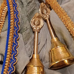 Könnte beinhalten: Zwei goldfarbene Glocken mit verzierten Griffen, die auf einer grauen Stoffoberfläche liegen. Die Griffe haben ein dekoratives Design mit einem erhabenen Gesicht.