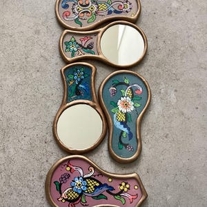 以下が含まれることがあります： 花柄の装飾が施された小さな手描きの鏡が5つ。鏡は涙型で、金色の枠があります。花柄は青、ピンク、黄色、緑の色合いです。