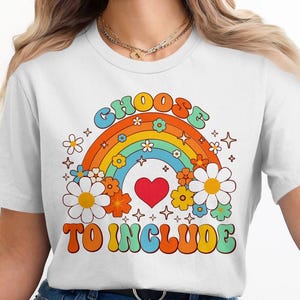 Puede incluir: Camiseta blanca con un gráfico de arcoíris y flores. El texto "Choose to Include" está impreso en una fuente retro.