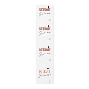 May include: A white rectangular table runner featuring a repeating pattern of the text DETROIT Genius in a salmon-colored serif font for the word DETROIT and a black cursive script for the word Genius, with small circular icons above and below.
