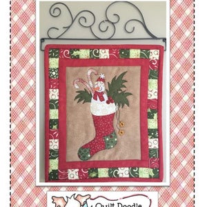 May include: A red and green patchwork quilt block with a snowman in a stocking. The quilt block is hanging on a black metal hook. The quilt block is titled "Jingle Snowman 145" and is a design by Quilt Doodle Designs.