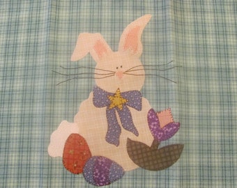 May include: A pink and white patchwork bunny with a blue bow and a yellow star. The bunny is holding a purple flower and is surrounded by three colorful eggs.