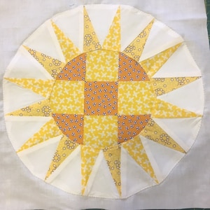 Puede incluir: Un fondo de tela blanco con un diseño de sol de patchwork amarillo y naranja. El sol está hecho de cuadrados y triángulos de tela.