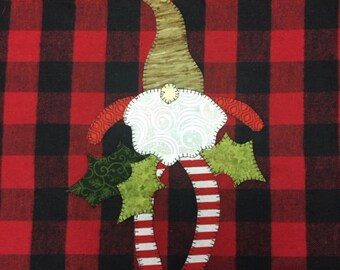 May include: A red and black plaid fabric background with a felt gnome applique. The gnome is wearing a white hat with a brown felt brim, a red and white striped shirt, and green holly leaves.