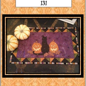 May include: A purple and black quilted table runner with a black cat and two jack-o'-lantern pumpkins. The runner has a black, white, and orange border. The text "It's a Black Cat Halloween 131" is at the top of the image. The text "Quilt Doodle Designs" is at the bottom of the image.