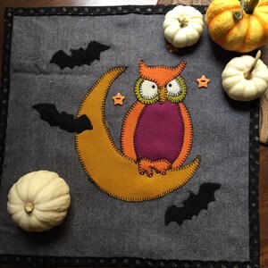 May include: A gray felt Halloween-themed wall hanging with an orange owl perched on a yellow crescent moon. There are three black bats and three white pumpkins on the gray background.
