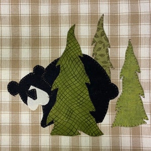 May include: A black bear peeking out from behind two green trees. The trees are made of fabric and have a stitched design. The bear is also made of fabric and has a white patch on its chest.