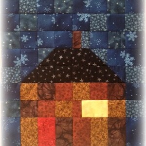 May include: A patchwork quilt block depicting a house with a chimney and a brown roof against a blue background with white snowflakes.
