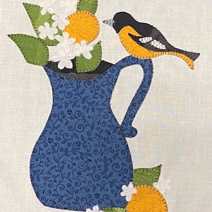May include: A blue pitcher with a floral pattern filled with white flowers, orange fruit, and green leaves. A black and orange bird sits on the rim of the pitcher.