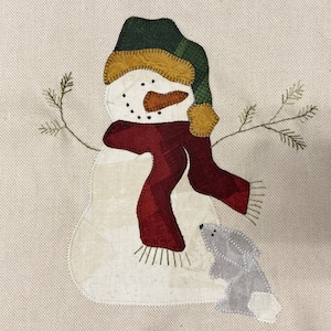 May include: A fabric snowman with a green and yellow hat, a burgundy scarf, and a carrot nose. The snowman has a small grey bunny friend. The background is a light beige colour, and there are two small branches on either side of the snowman.